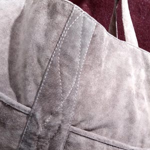 May include: A gray suede tote bag with a stitched design on the front. The bag has two straps.
