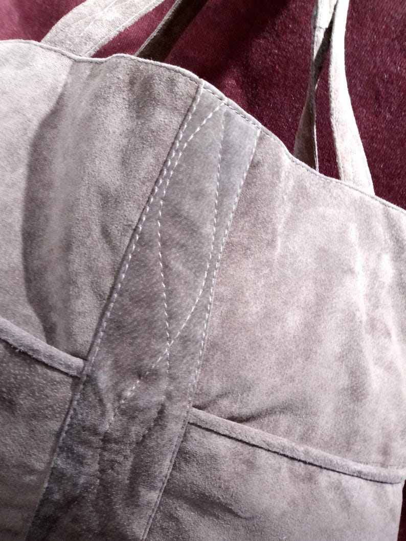 May include: A gray suede tote bag with a stitched design on the front. The bag has two straps.