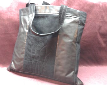May include: A black tote bag featuring a central panel with a stitched leaf pattern, flanked by smooth black side panels. The bag includes two black fabric handles and a horizontal top trim, constructed from a combination of textured and smooth materials.
