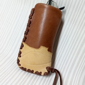 May include: A brown leather glasses case with a tan leather patch and a braided leather strap. The case has a flap that closes with a snap.