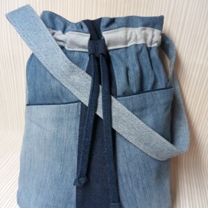 May include: A denim bucket bag with a light blue shoulder strap and a dark blue drawstring closure. The bag features a front pocket and is made from various shades of blue denim.