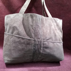 May include: A gray suede tote bag with two top handles. The bag has a large pocket on the front and a smaller pocket on the back.