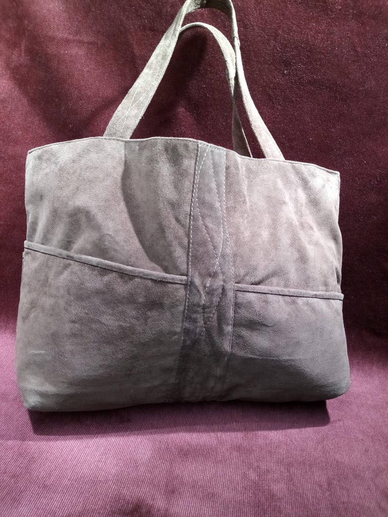May include: A gray suede tote bag with two top handles. The bag has a large pocket on the front and a smaller pocket on the back.