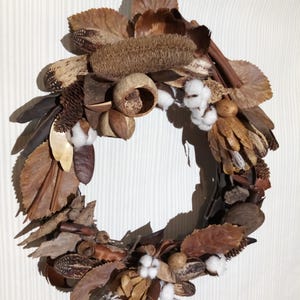 May include: A circular wreath made from dried leaves, seed pods, cotton bolls, and other natural elements in shades of brown, beige, and white. The wreath is suspended by a black ribbon, suitable for seasonal home decoration.
