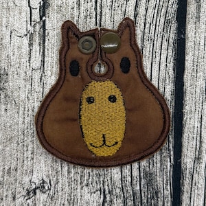 Op de afbeelding: Een bruine stoffen patch in de vorm van een capybarakop met een gele geborduurde snuit, zwarte gestikte ogen en een klein mondje. De patch is voorzien van twee donkerbruine plastic drukknopen aan de bovenkant om rond een object te bevestigen.