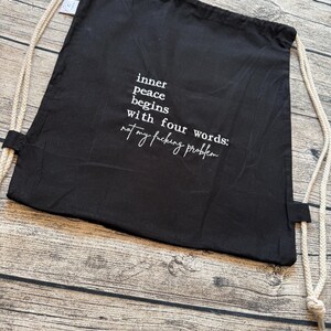 Puede incluir: Mochila negra con cord&oacute;n y texto blanco: "la paz interior comienza con cuatro palabras: no es mi puto problema". La bolsa tiene correas de cuerda beige y est&aacute; sobre una superficie de madera.