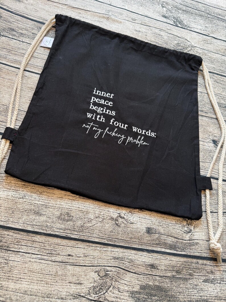 Puede incluir: Mochila negra con cord&oacute;n y texto blanco: "la paz interior comienza con cuatro palabras: no es mi puto problema". La bolsa tiene correas de cuerda beige y est&aacute; sobre una superficie de madera.