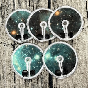 May include: Five circular fabric pads featuring a galaxy print with stars and nebulae in shades of teal, black, and orange. Each pad has a white stitched border, a keyhole-shaped cutout, and two black plastic snap fasteners at the bottom edge.