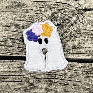 Op de afbeelding: Een witte geborduurde spookpatch met twee zwarte ovale ogen en een bloemenkrans bestaande uit drie bloemen in paars, lichtroze en geel. Het spookje heeft een centrale ronde uitsparing en twee kleine witte ronde drukknopen aan de onderkant.