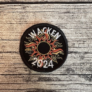 Pode incluir: Um patch circular preto com a palavra "WACKEN" em branco, arqueada na parte superior. Abaixo, um desenho estilizado de sol em laranja e amarelo circunda um centro preto. O ano "2024" está escrito em branco na parte inferior. O patch tem uma borda preta.