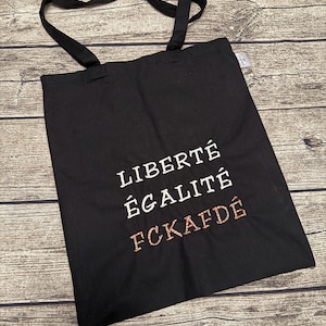 Puede incluir: Bolso tote negro con asas largas. La bolsa presenta las palabras "LIBERTÉ ÉGALITÉ FCKAFDÉ" bordadas en hilo blanco y rojo. La bolsa está hecha de una tela resistente y es adecuada para transportar varios artículos.