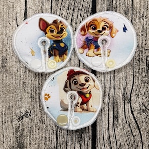 Könnte beinhalten: Drei runde Stoffaufnäher mit gestickten Cartoon-Hundefiguren. Jeder Aufnäher hat einen Knopfverschluss und eine schlüssellochförmige Öffnung. Die Hunde sind in verschiedenen Posen zu sehen, tragen Uniformen und lächeln. Der Hintergrund ist hellblau und weiß.