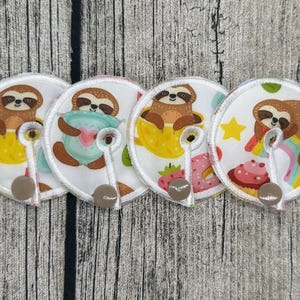 Könnte beinhalten: Vier kreisförmige Stilleinlagen aus Stoff mit weißem Hintergrund und bunten Faultier-Illustrationen, darunter Faultiere in gelben Tassen, mit Herzen und Regenbögen, jeweils mit einer kleinen weißen Lasche und einem braunen Druckknopf.