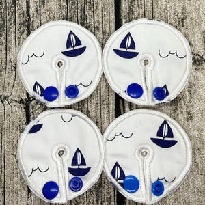 Könnte beinhalten: Vier kreisförmige weiße Stoffpads mit einem dunkelblauen Segelboot- und Wellenmuster. Jedes Pad hat eine mittige Öffnung mit einem Schlitz bis zum Rand, der mit zwei leuchtend blauen Kunststoff-Druckknöpfen zum Verschließen versehen ist.