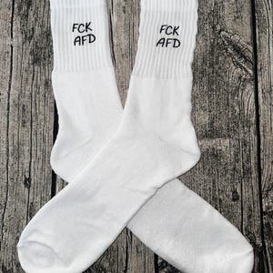 Może przedstawiać: Para białych skarpet sportowych z napisem "FCK AFD" wyhaftowanym na czarno na prążkowanym mankiecie. Skarpetki wykonane są z miękkiego materiału i prezentowane na drewnianej powierzchni.