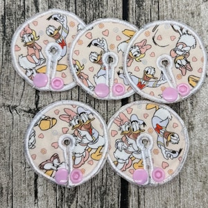 Könnte beinhalten: Fünf kreisförmige Stoffabdeckungen mit einem wiederkehrenden Muster von Donald und Daisy Duck auf hellpfirsichfarbenem Hintergrund mit kleinen rosa Herzen und Blumen. Jede Abdeckung hat ein Mittelloch, weiße Nähte und zwei kleine rosa Druckknöpfe.