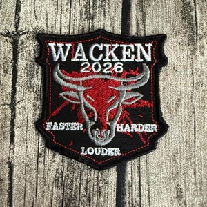 May include: A shield-shaped embroidered patch with a black background and red stitching. It features a silver bull head graphic, the text WACKEN 2026 at the top, and the words FASTER HARDER LOUDER arranged around the bull in white thread.