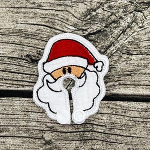 Op de afbeelding: Een witte stoffen speenkoord met een geborduurd gezicht van de Kerstman, voorzien van een rode muts, een beige huidskleur en een witte baard. Het ontwerp heeft een ronde uitsparing in het midden en twee witte plastic drukknopen onderaan.