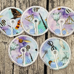 Könnte beinhalten: Fünf kreisförmige Kabelhalter aus Stoff mit einem Aquarell-Druck verschiedener Fantasy-Motive, darunter ein sprechender Hut, ein Zauberstab, ein goldener Schnatz, eine Brille und Schlüssel. Weiße Kanten und zwei hellblaue Kunststoff-Druckknöpfe.