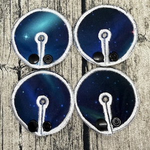 Peut inclure: Quatre coussinets en tissu circulaires avec un imprimé galaxie bleu foncé et sarcelle, bordés de broderies blanches. Chaque coussinet présente un motif brodé en forme de trou de serrure et deux boutons-pression noirs en plastique.