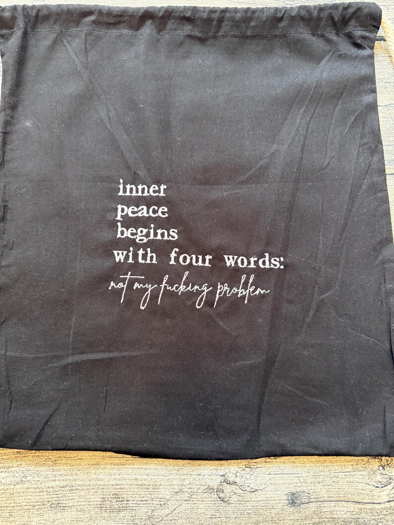 Puede incluir: Bolsa negra con cord&oacute;n y texto blanco: "la paz interior comienza con cuatro palabras: no es mi puto problema."