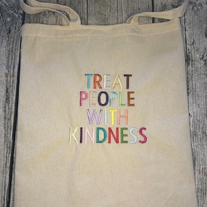 Könnte beinhalten: Beige Canvas-Tragetasche mit zwei Griffen. Der Beutel hat den gestickten Schriftzug "TREAT PEOPLE WITH KINDNESS" in bunten Blockbuchstaben. Die Tasche besteht aus einem robusten Stoff und eignet sich zum Tragen verschiedener Gegenstände.