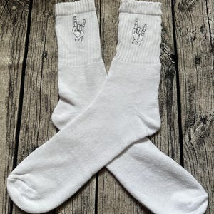 May include: A pair of white crew socks with a black embroidered hand gesture on the cuff. The socks are made of a soft, ribbed material and are displayed on a wooden surface. The hand gesture is a symbol of rock and roll.