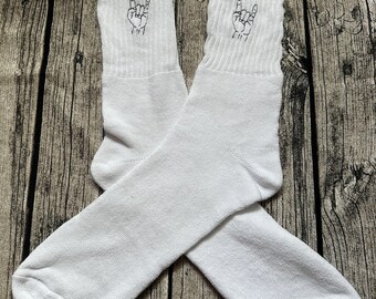 May include: A pair of white crew socks with a black embroidered hand gesture on the cuff. The socks are made of a soft, ribbed material and are displayed on a wooden surface. The hand gesture is a symbol of rock and roll.