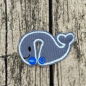 Op de afbeelding: Een grijze walvisvormige stoffen applicatie met een wafelstructuur, voorzien van een lichtblauw geborduurde omlijning, een klein zwart gestikt oog en mond, en twee felblauwe plastic drukknopen op het onderste gedeelte van het lichaam.