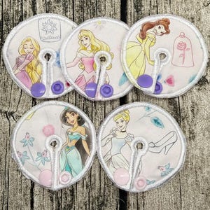 Könnte beinhalten: Fünf kreisförmige Stoffpads mit Zeichentrick-Prinzessinnen-Illustrationen, darunter Rapunzel, Aurora, Belle, Jasmin und Aschenputtel. Jedes Pad hat einen mittigen Schlitz, weiß vernähte Ränder und zwei kleine Kunststoff-Druckknöpfe in Lila oder Rosa.