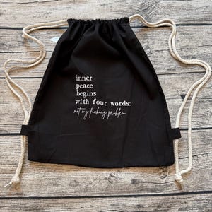 Puede incluir: Mochila negra con cord&oacute;n y texto blanco: "la paz interior comienza con cuatro palabras: not my fucking problem". La bolsa tiene correas de cuerda blancas. El fondo es una superficie de madera. La bolsa est&aacute; hecha de tela oscura.