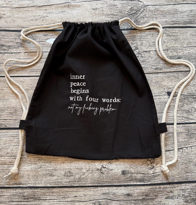 Puede incluir: Mochila negra con cord&oacute;n y texto blanco: "la paz interior comienza con cuatro palabras: not my fucking problem". La bolsa tiene correas de cuerda blancas. El fondo es una superficie de madera. La bolsa est&aacute; hecha de tela oscura.