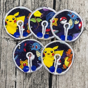 Op de afbeelding: Vijf ronde stoffen patches met geborduurde stripfiguren op een sterrenachtergrond. Elke patch heeft een witte rand en een blauwe knoopsluiting. Pikachu en Charmander zijn afgebeeld.