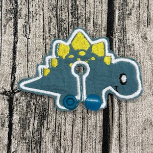 Op de afbeelding: Een blauwgroene dinosaurusvormige stoffen patch met gele geborduurde platen op de rug. De patch heeft een witte gestikte omlijning, een kleine ronde uitsparing in het midden en twee blauwe plastic drukknopen aan de onderkant.
