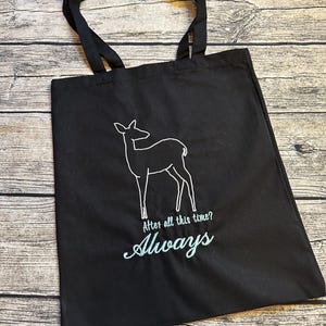Op de afbeelding: Zwarte draagtas met een witte omtrek van een hert en de tekst "After all this time? Always" in lichtblauw script. De tas heeft twee zwarte handvatten en is gemaakt van een stevige stof.
