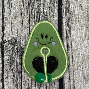 Könnte beinhalten: Ein grüner Kabel-Organizer aus Stoff in Avocadoform mit einem gestickten lächelnden Gesicht, violetten Wangen und einem schwarzen herzförmigen Kern. Das Design umfasst einen mittigen runden Ausschnitt und einen kleinen grünen Druckknopf am unteren Rand.