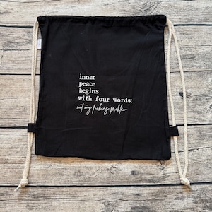 Könnte beinhalten: Schwarzer Turnbeutel mit weißem Text: "inner peace begins with four words: not my fucking problem". Der Beutel hat cremefarbene Kordelzüge und wird auf einer Holzoberfläche präsentiert.