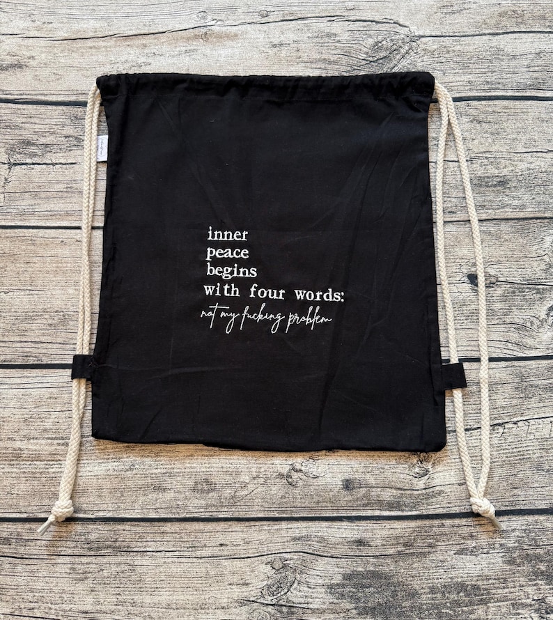 Puede incluir: Bolsa con cord&oacute;n negra con texto blanco: "inner peace begins with four words: not my fucking problem". La bolsa tiene cordones color crema y se muestra sobre una superficie de madera.