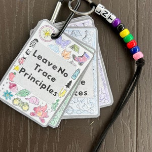 May include: A set of colorful cards with illustrations of outdoor activities and the text "Leave No Trace Principles". The cards are attached to a black carabiner with a beaded keychain.