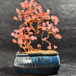 May include: A handcrafted bonsai tree sculpture with a copper wire trunk and branches, adorned with pink crystal leaves. The tree sits in a glazed blue ceramic pot with a textured surface, creating a unique decorative piece.