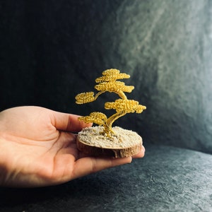 Könnte beinhalten: Eine goldene Drahtskulptur eines Bonsai-Baums auf einem Holzsockel mit hellbrauner Rinde und weißem Sand.