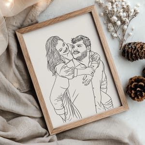 Peut inclure: Une impression encadrée d'art linéaire représente un homme portant une femme sur son dos. L'illustration en noir et blanc est placée sur un fond crème dans un cadre en bois brun clair. L'œuvre capture un moment d'affection et de joie.