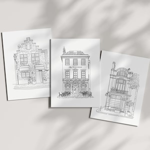Op de afbeelding: Drie witte rechthoekige kunstdrukken met zwarte lijntekeningen van gebouwen in Europese stijl. De middelste print toont een gebouw met het label The Huntsman, terwijl de linker print een winkelpui toont met de tekst Chocolate en Jene.