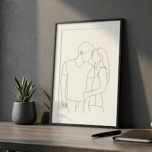 May include: A framed line art print showing a couple in an embrace, kissing. The minimalist design uses black lines on a cream background. The print is in a black frame, displayed on a wooden surface with a potted plant.