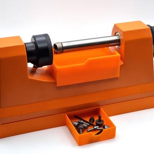 Peut inclure: Un tour d'établi orange doté d'une broche métallique horizontale, d'un mandrin noir et d'une poignée argentée. Un petit plateau orange fixé à l'avant contient plusieurs petits composants métalliques, notamment des goupilles et des vis.