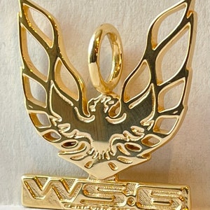 May include: A gold pendant depicting a bird with open wings. The pendant has a loop for a chain and a rectangular base with the text "WSE PERFORMANCE GARAGE". The design is detailed and intricate, with a polished finish.