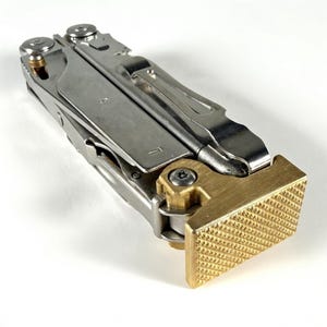 May include: A multi-tool with a silver metal body and a brass-colored textured end. The tool has multiple components and a clip. The brass end has a rectangular shape with a grid-like pattern.