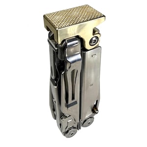 May include: A multi-tool with a brass-colored textured top and a stainless steel body. The tool is closed, showing its various components and a small screw. The design is compact and appears durable.