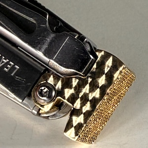 May include: Close-up of a multi-tool with a gold-colored, diamond-patterned hammer head. The tool is made of silver-colored metal and has the word "LEA" partially visible. The hammer head has a textured surface.