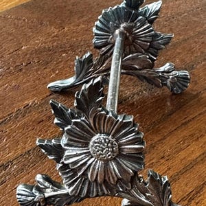 May include: A silver-toned metal stand featuring two floral motifs with detailed petals and leaves. The structure consists of two identical flower-shaped supports connected by a central horizontal bar, designed to hold items in an upright position.